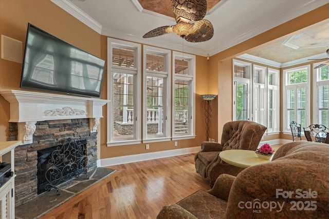 living room featuring a ceiling fan, a stone fireplace, baseboards, wood finished floors, and crown molding
