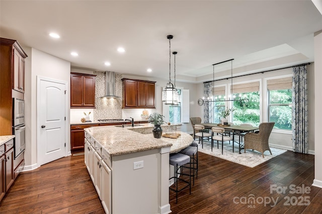 kitchen featuring wall chimney exhaust hood, a sink, dark wood-style floors, light stone counters, and an island with sink