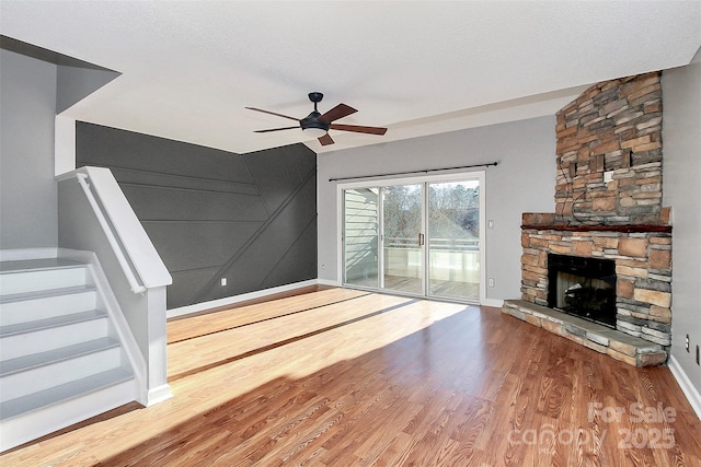 unfurnished living room featuring stairs, wood finished floors, a stone fireplace, baseboards, and a ceiling fan