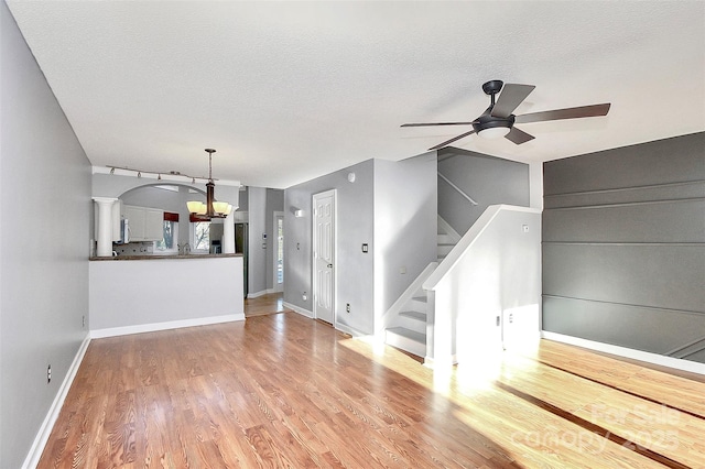 unfurnished living room featuring baseboards, stairway, light wood-type flooring, a textured ceiling, and ceiling fan with notable chandelier
