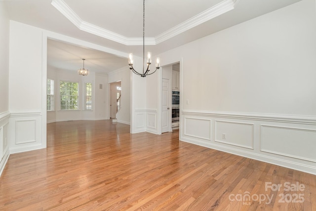 unfurnished dining area with light wood-style floors, a raised ceiling, an inviting chandelier, ornamental molding, and a decorative wall