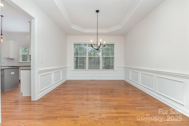 unfurnished dining area with an inviting chandelier, ornamental molding, a raised ceiling, a wainscoted wall, and light wood-style flooring