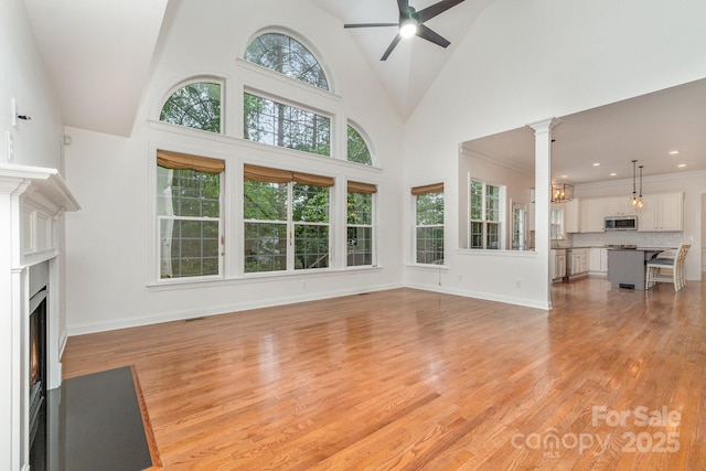 unfurnished living room with light wood finished floors, high vaulted ceiling, a ceiling fan, a fireplace with flush hearth, and decorative columns