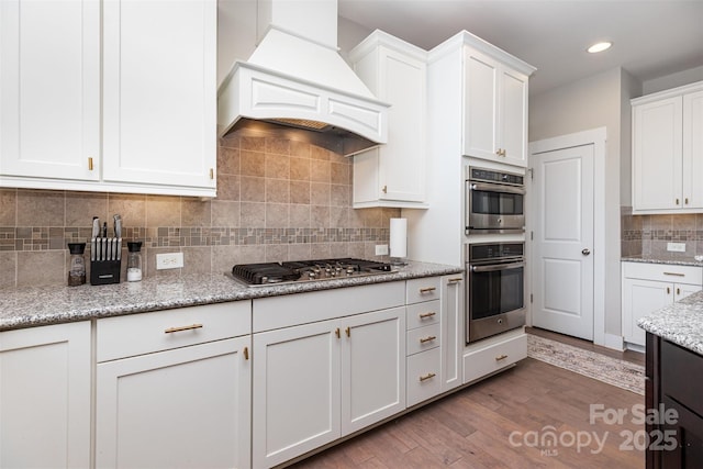 kitchen featuring custom exhaust hood, stainless steel appliances, white cabinetry, decorative backsplash, and wood finished floors