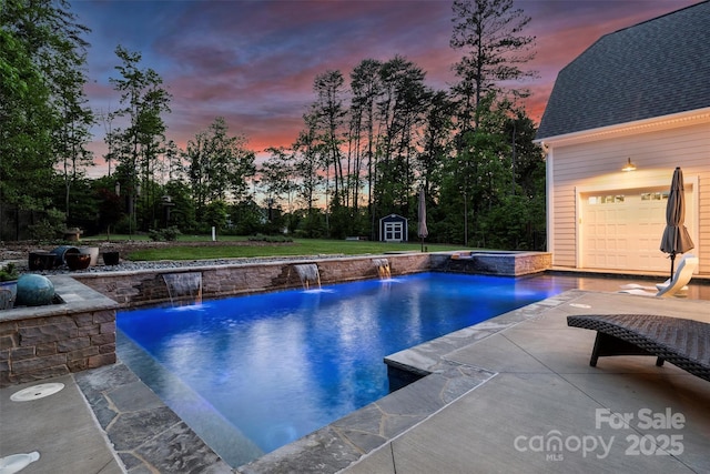 pool at dusk featuring a pool with connected hot tub, a storage unit, a patio, an outbuilding, and a lawn