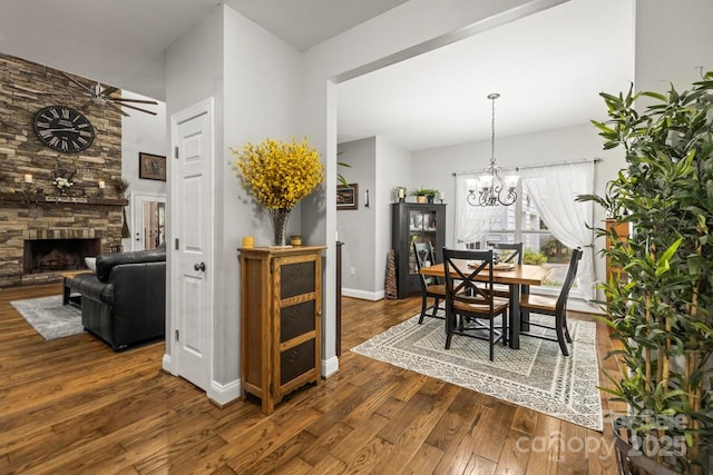 dining room with dark wood-style floors, a stone fireplace, a chandelier, ceiling fan, and baseboards