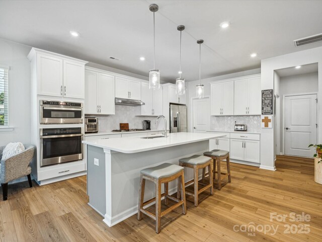 kitchen featuring appliances with stainless steel finishes, light wood-style floors, white cabinetry, tasteful backsplash, and under cabinet range hood