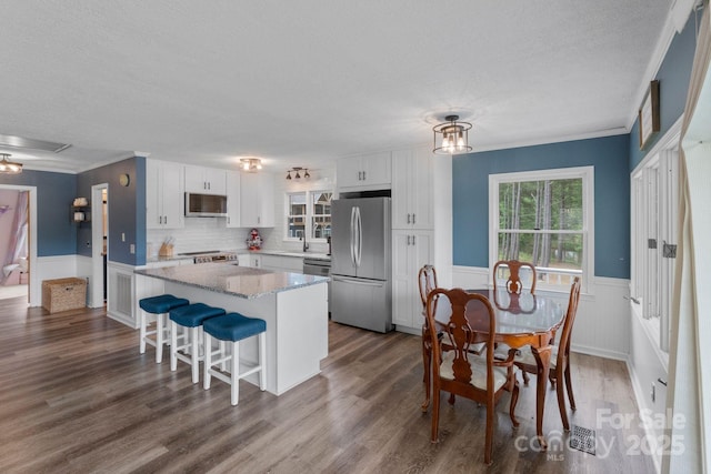 kitchen with appliances with stainless steel finishes, a textured ceiling, a kitchen island, white cabinetry, and wainscoting