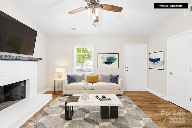living room with a ceiling fan, visible vents, a brick fireplace, wood finished floors, and baseboards