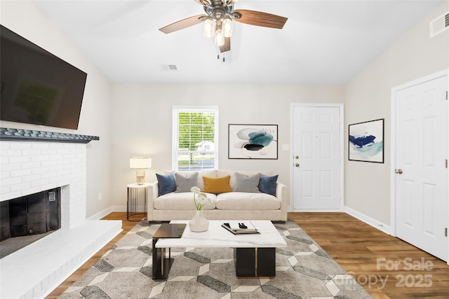 living area featuring a ceiling fan, visible vents, a brick fireplace, wood finished floors, and baseboards
