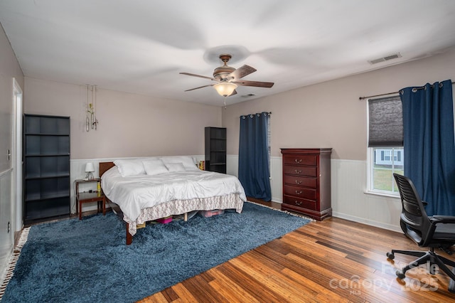 bedroom with a ceiling fan, visible vents, wood finished floors, and a wainscoted wall