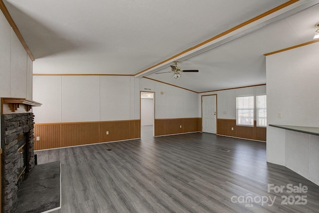 unfurnished living room featuring a stone fireplace, dark wood-style floors, vaulted ceiling with beams, a wainscoted wall, and a ceiling fan