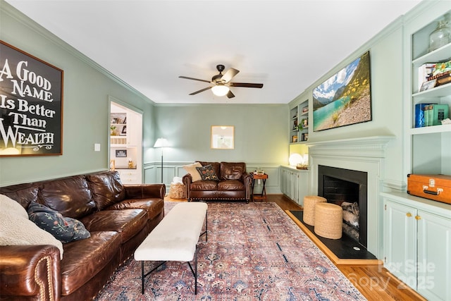 living area with ceiling fan, wainscoting, a fireplace, built in features, and wood finished floors