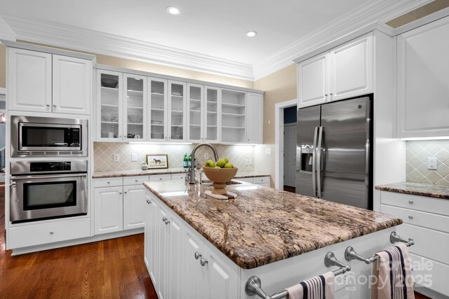 kitchen with glass insert cabinets, an island with sink, tasteful backsplash, light stone counters, and appliances with stainless steel finishes