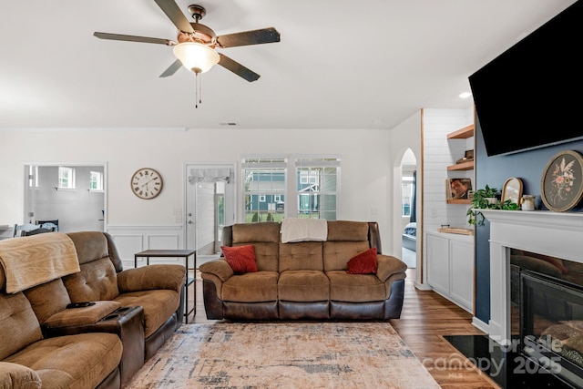 living area featuring plenty of natural light, a ceiling fan, a fireplace with flush hearth, arched walkways, and wood finished floors