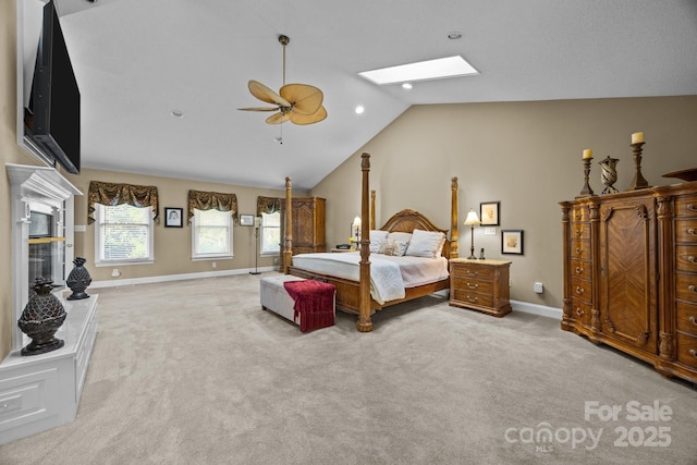 bedroom featuring ceiling fan, light colored carpet, a fireplace with raised hearth, baseboards, and lofted ceiling with skylight