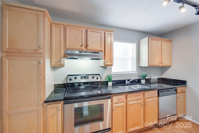 kitchen featuring stainless steel appliances, light brown cabinetry, under cabinet range hood, a sink, and a textured ceiling