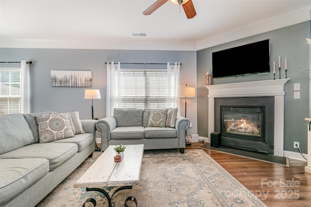 living area with ceiling fan, visible vents, crown molding, a fireplace with flush hearth, and wood finished floors