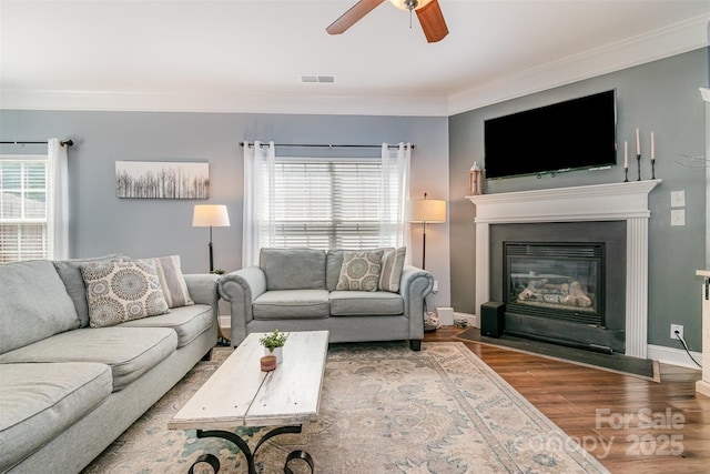 living room with visible vents, ornamental molding, a glass covered fireplace, wood finished floors, and ceiling fan