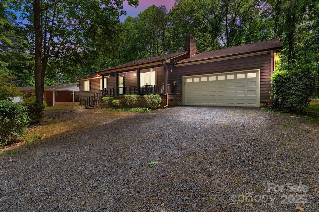 view of front of home featuring covered porch, an attached garage, a chimney, and gravel driveway