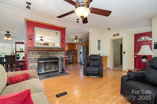 living room featuring a textured ceiling, visible vents, a stone fireplace, ceiling fan, and light wood-style floors