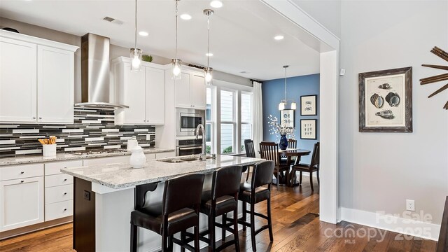 kitchen featuring decorative backsplash, a kitchen island with sink, wall chimney range hood, white cabinetry, and appliances with stainless steel finishes