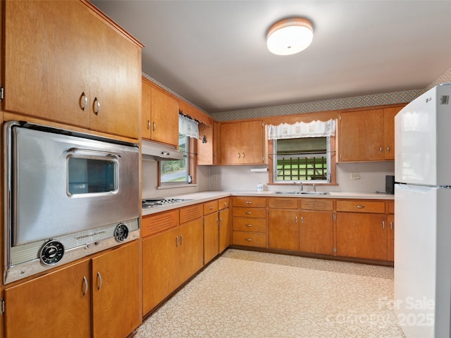 kitchen with a sink, light countertops, freestanding refrigerator, under cabinet range hood, and stovetop