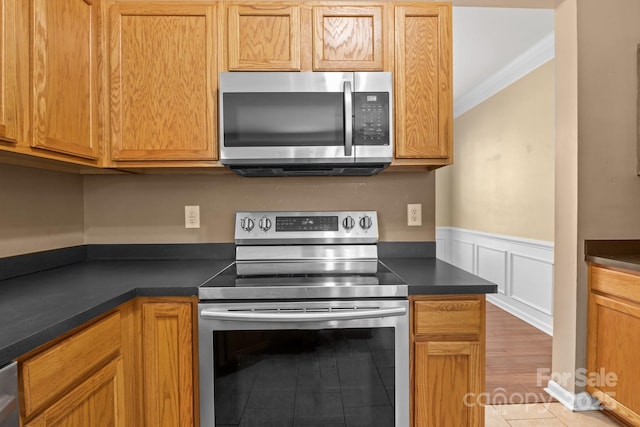 kitchen with crown molding, a decorative wall, dark countertops, a wainscoted wall, and stainless steel appliances