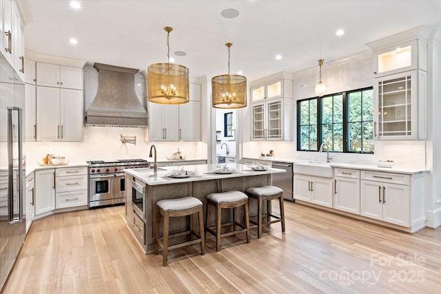 kitchen featuring a sink, light wood finished floors, an island with sink, premium range hood, and stainless steel appliances