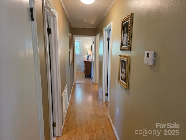 corridor with a textured ceiling, visible vents, light wood-type flooring, baseboards, and ornamental molding