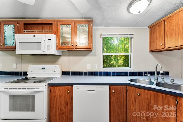 kitchen featuring brown cabinets, a sink, white appliances, a textured ceiling, and glass insert cabinets