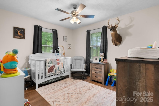 bedroom featuring a nursery area, a ceiling fan, multiple windows, and wood finished floors