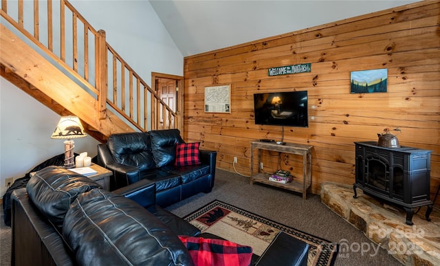 living room featuring carpet flooring, high vaulted ceiling, a wood stove, wooden walls, and stairs