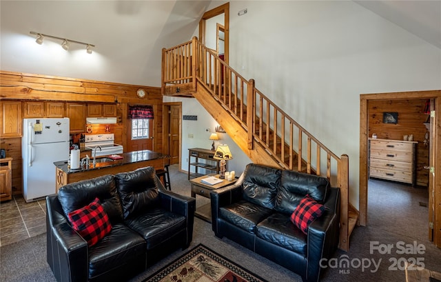 living room with stairs, wood walls, dark colored carpet, and high vaulted ceiling