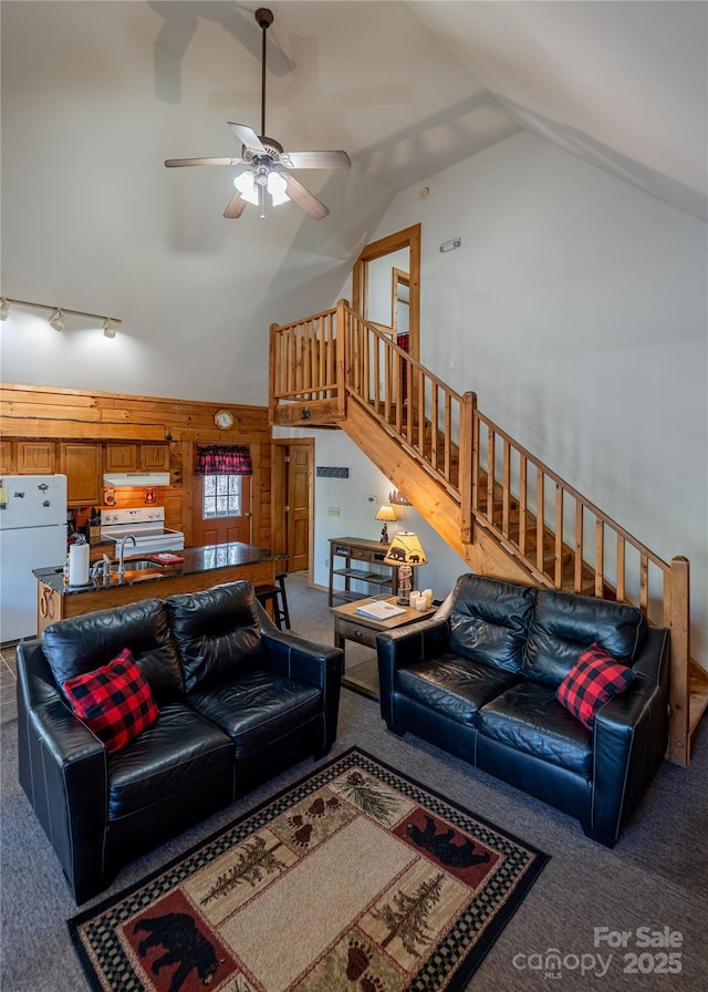 living room featuring wood walls, stairs, high vaulted ceiling, carpet floors, and a ceiling fan