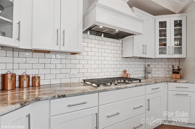 kitchen with light stone countertops, custom range hood, white cabinetry, and backsplash
