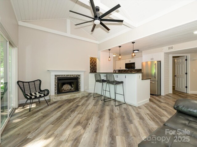 living area with ceiling fan, visible vents, a tiled fireplace, light wood-type flooring, and vaulted ceiling with beams
