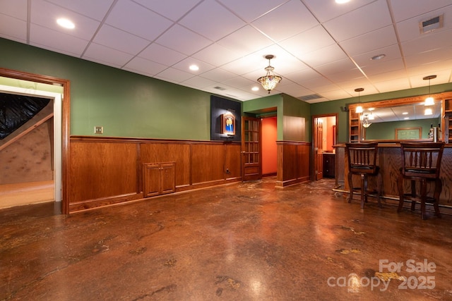 bar featuring a paneled ceiling, hanging light fixtures, concrete flooring, a wainscoted wall, and indoor bar