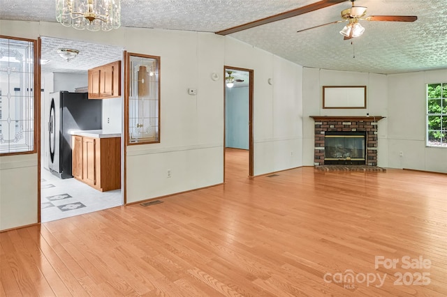 unfurnished living room featuring a brick fireplace, light wood-style floors, vaulted ceiling with beams, a textured ceiling, and ceiling fan with notable chandelier
