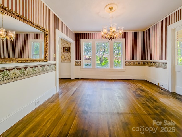unfurnished dining area featuring a chandelier, healthy amount of natural light, wood finished floors, wallpapered walls, and ornamental molding