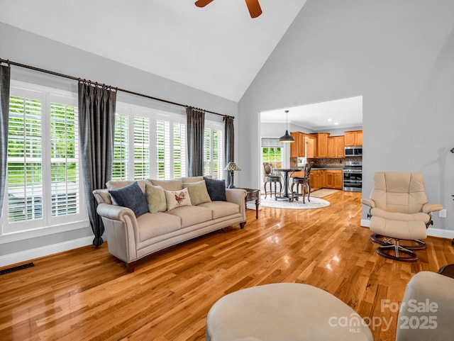 living room featuring baseboards, light wood-style flooring, ceiling fan, and high vaulted ceiling