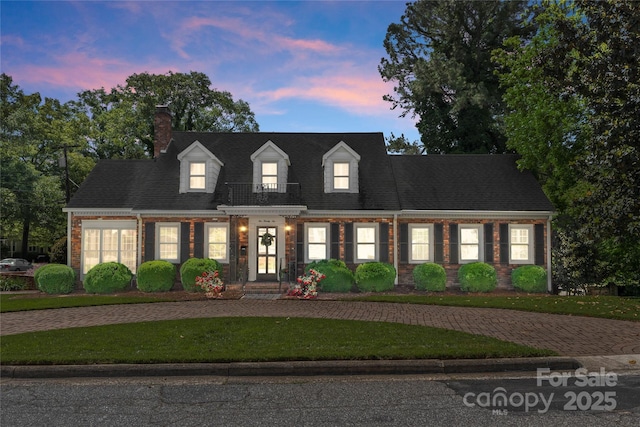 new england style home featuring brick siding, a front yard, a chimney, a balcony, and curved driveway