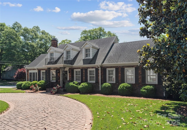new england style home with a balcony, brick siding, a chimney, a front yard, and curved driveway