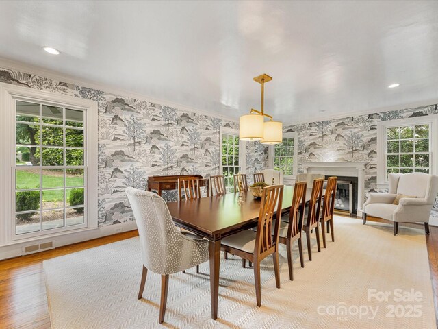 dining area featuring wallpapered walls, light wood-style floors, healthy amount of natural light, ornamental molding, and a high end fireplace