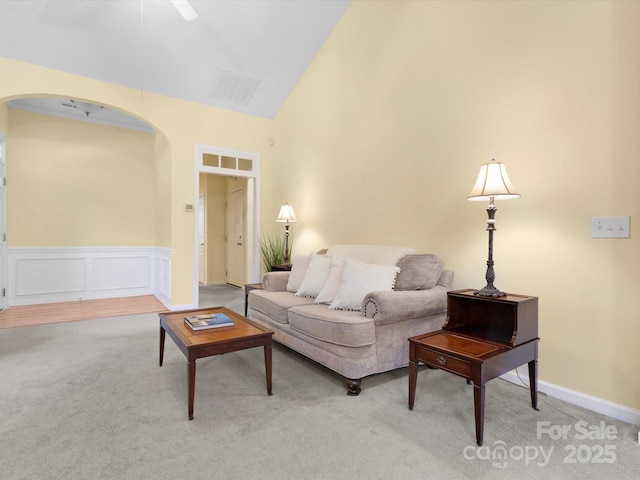 carpeted living room with ceiling fan, visible vents, arched walkways, vaulted ceiling, and wainscoting