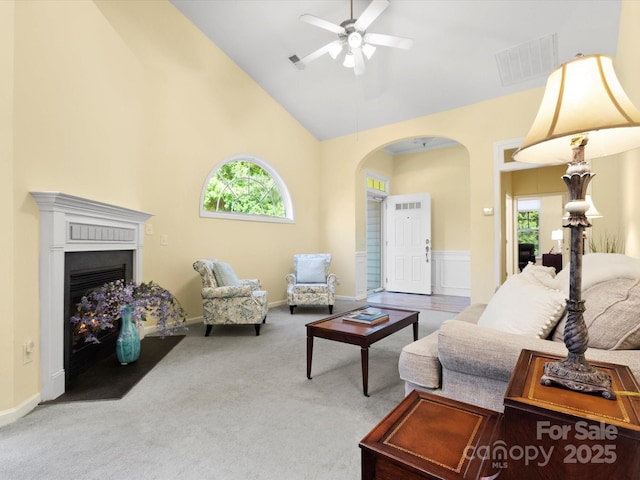 carpeted living area with visible vents, a fireplace with flush hearth, ceiling fan, high vaulted ceiling, and arched walkways