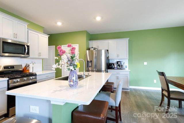 kitchen featuring wood finished floors, an island with sink, a sink, stainless steel appliances, and backsplash