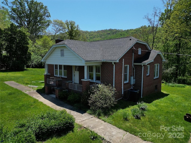 view of front of home featuring covered porch, a forest view, roof with shingles, brick siding, and a front lawn