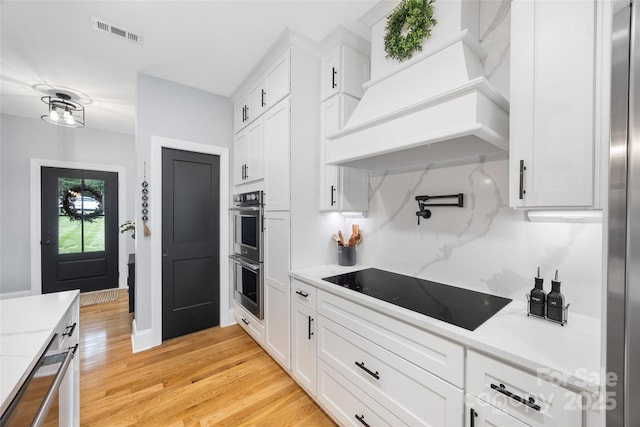 184 Quiet Cove Rd, Mooresville, NC 28117 - kitchen with black electric stovetop, premium range hood, double oven, white cabinetry, and light wood-type flooring