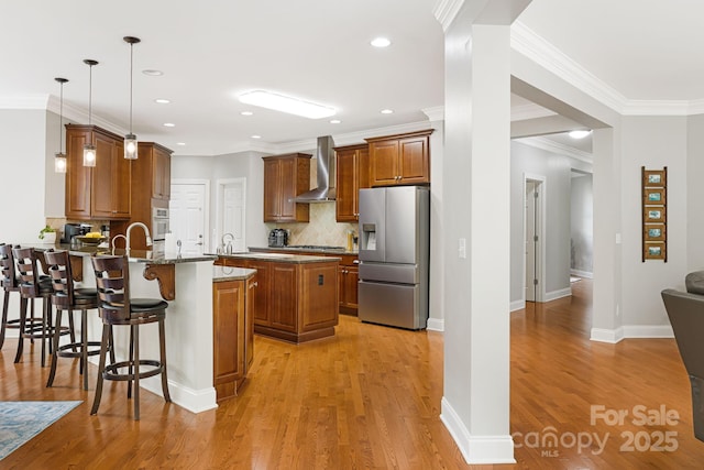 kitchen with a peninsula, brown cabinetry, wall chimney exhaust hood, a kitchen bar, and stainless steel fridge